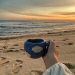 holding blue coffee cup on the beach with sunset view